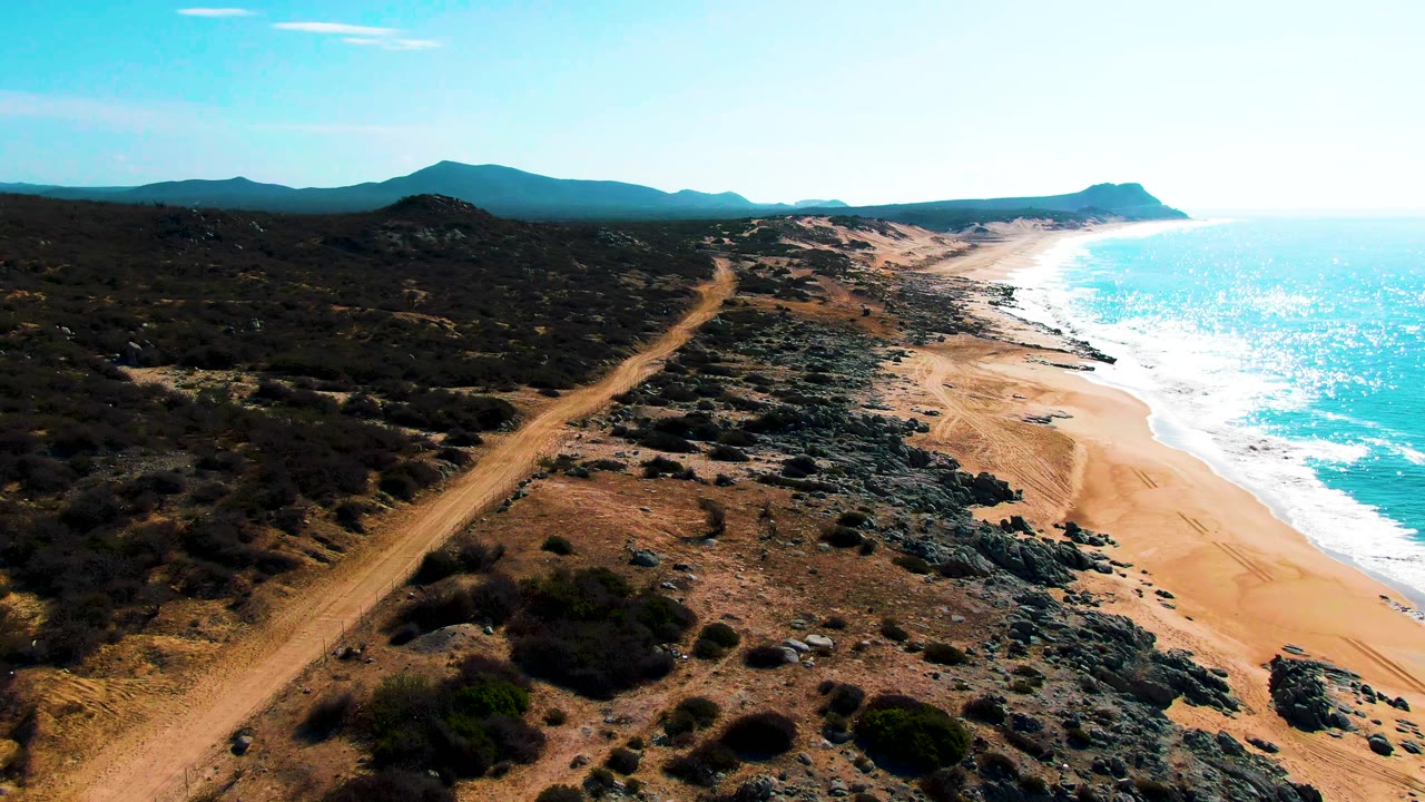 Aerial frame over Punta Buzzards with the dirt road cutting diagonally through scrubland, dark rocks above the sand, white surf along the beach, hazy hills, and no visible people.