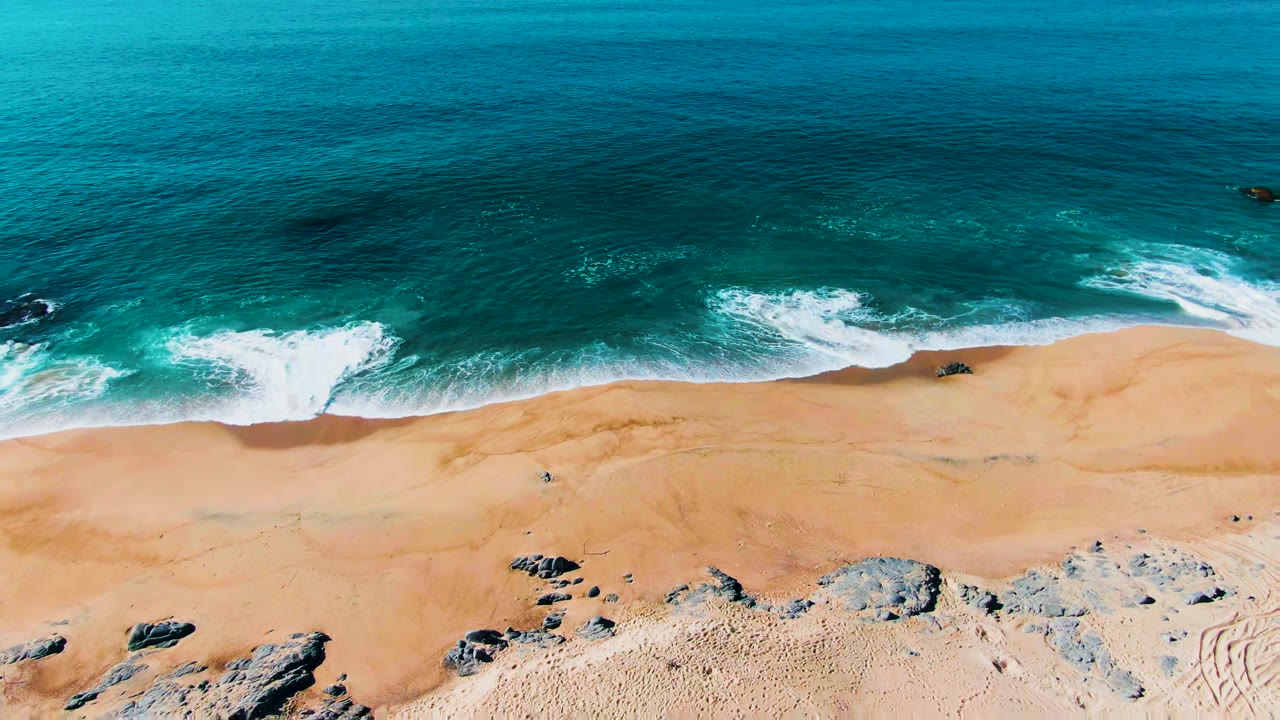 Overhead view at Punta Buzzards, Baja California Sur, showing turquoise ocean, white foam, sandy beach, scattered dark rocks, and no visible people at the start of the clip.