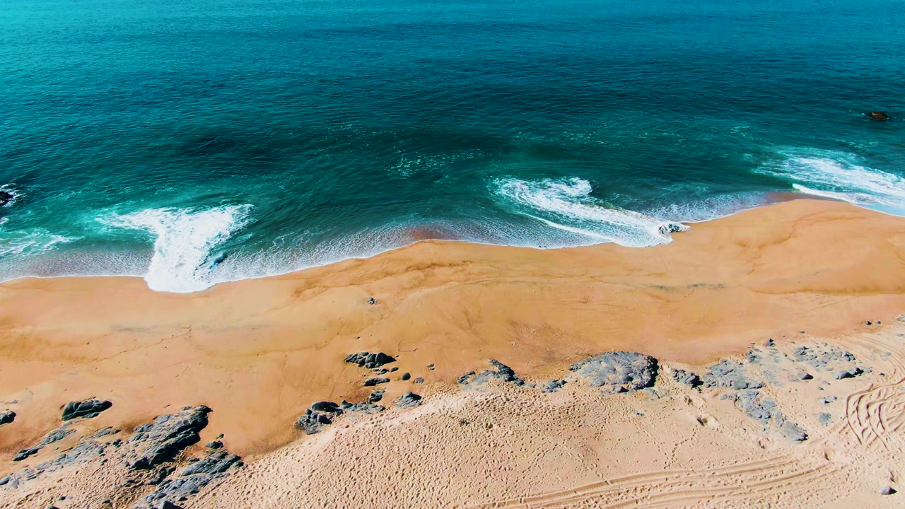 At 0:02, foamy water curls along the Punta Buzzards sand, with dark rocks near the lower edge, faint track marks to the right, and no visible people.