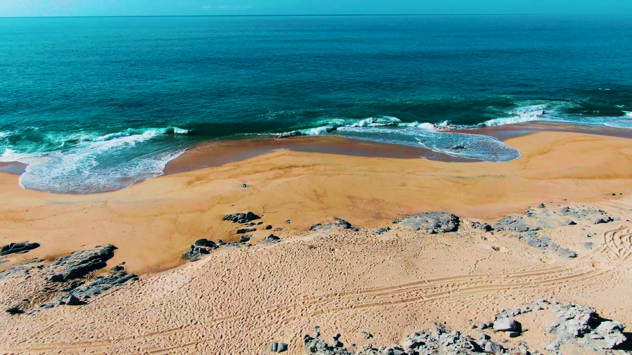 Aerial frame at Punta Buzzards, Baja California Sur showing turquoise ocean, a line of white surf, tan beach, scattered rocks, tire tracks, and no visible people.