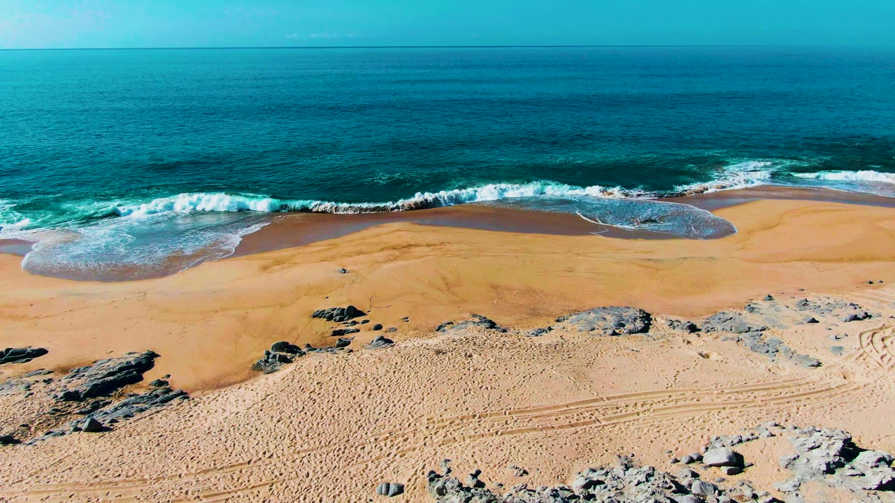 Aerial view at Punta Buzzards with the surf line shifted slightly along the wet beach, pale rocks in the lower frame, open ocean beyond, and no visible people.