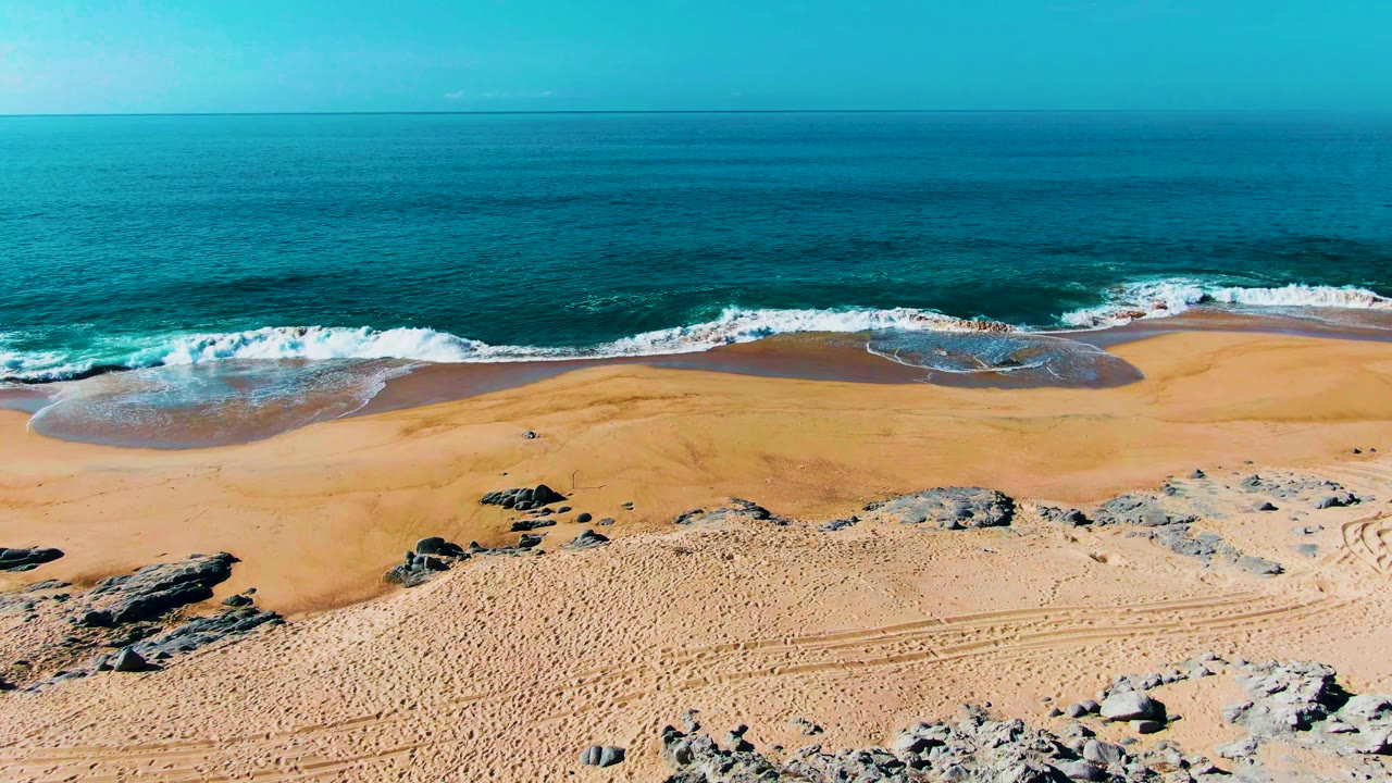 At Punta Buzzards, white foam stretches farther across the wet sand while rocky outcrops and beach tracks remain visible in the foreground, with no visible people.