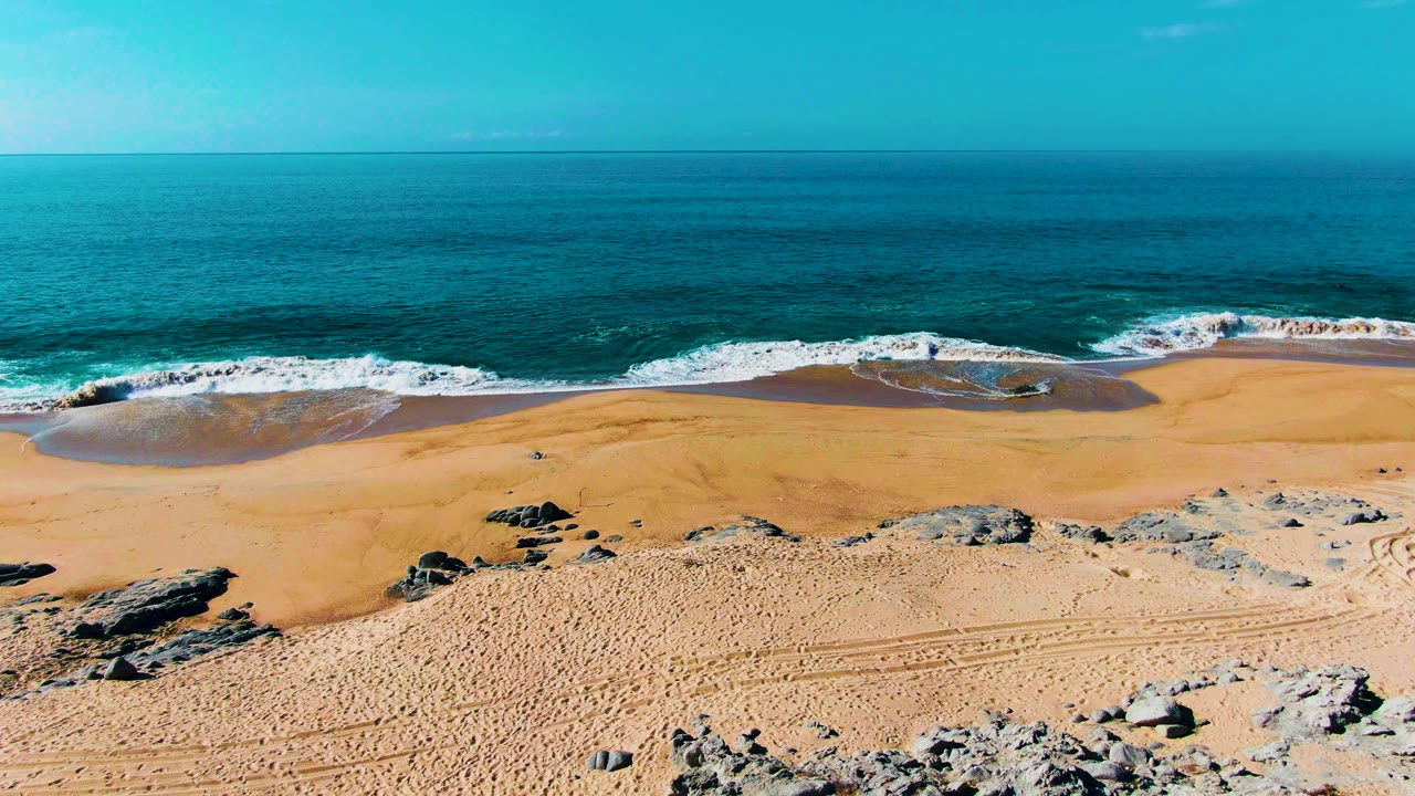 Elevated beach view with receding foam, sandy shore, rocks, and blue ocean.