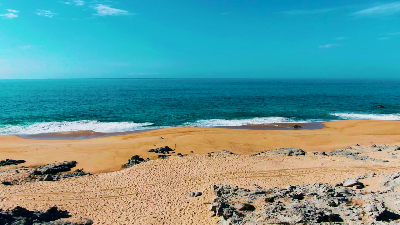 The Punta Buzzards shoreline fills the frame with tan sand, dark rocks near the lower edge, thin white breakers, and open blue water, with no visible people.