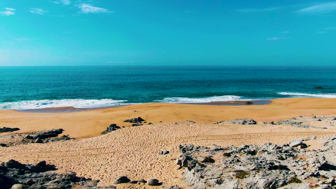 Rocks spread across the lower right of the Punta Buzzards view while a broad sandy beach meets turquoise water and white foam, with no visible people.