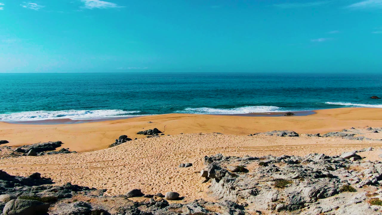 The elevated Punta Buzzards view shows empty sand, scattered black rocks, a low line of breaking waves, and a clear blue sky, with no visible people.