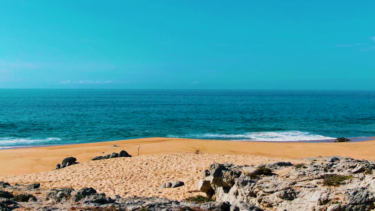 Buzzards Beach shoreline in Punta Buzzards, Mexico, seen over pale rocks and dry sand, with white surf lines on blue water and no visible people.
