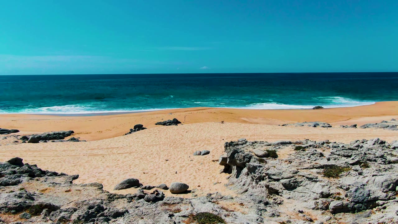 Elevated view of Buzzards Beach in Punta Buzzards, Baja California Sur, with pale sand, dark rocks in the foreground, turquoise surf, a blue horizon, and no visible people.