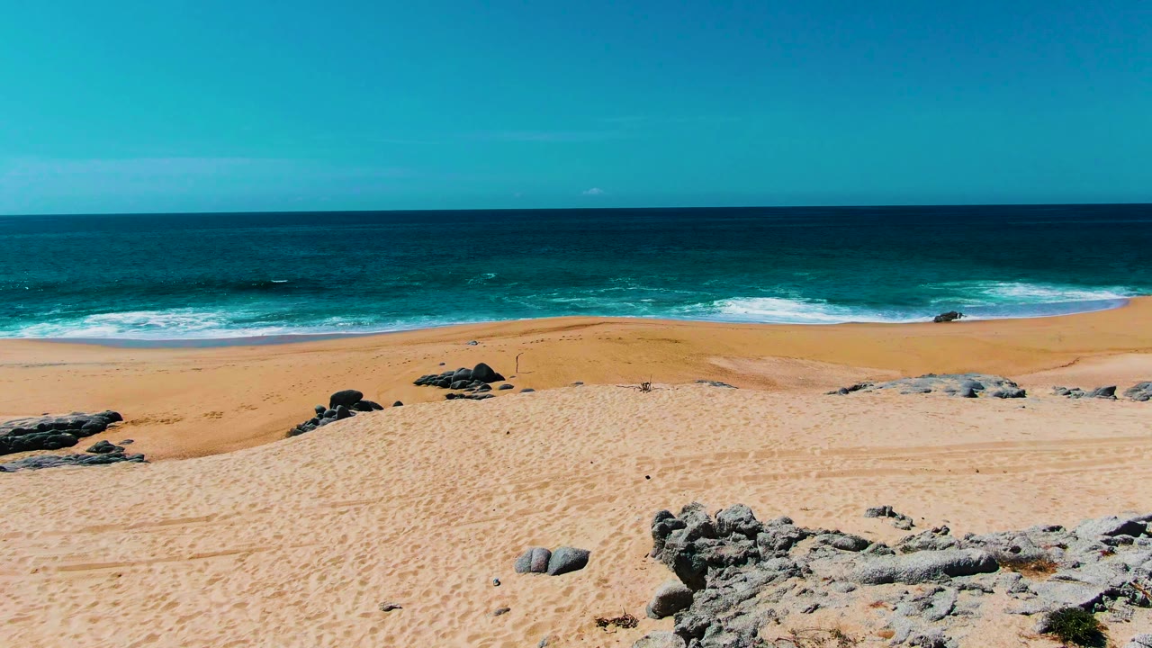 A wider section of Buzzards Beach shows scattered dark rocks on golden sand, white wave foam near shore, deep blue water toward the horizon, and no visible people.