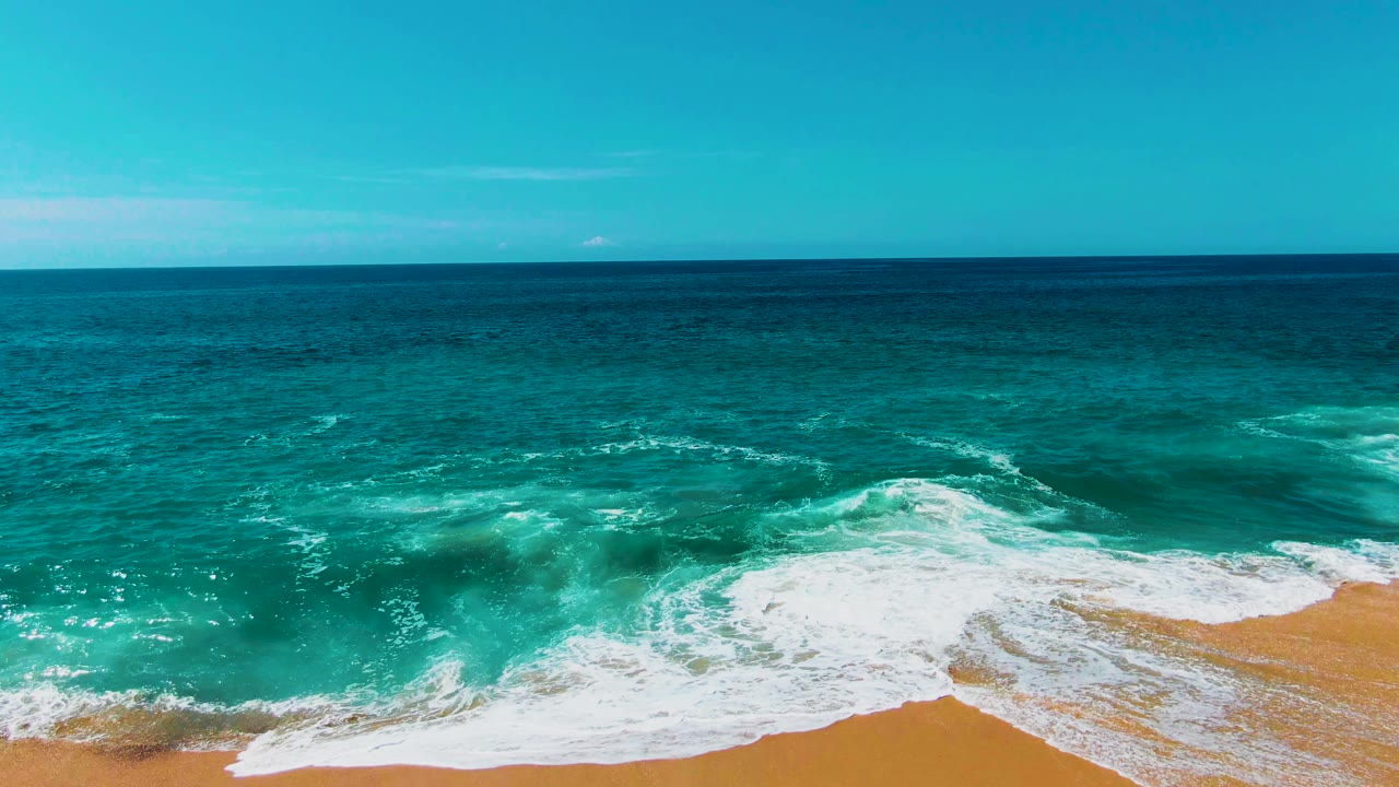 At Punta Buzzards in Baja California Sur, turquoise surf pushes white foam across the sandy lower edge while deeper blue ocean and clear sky fill the frame, with no visible people.