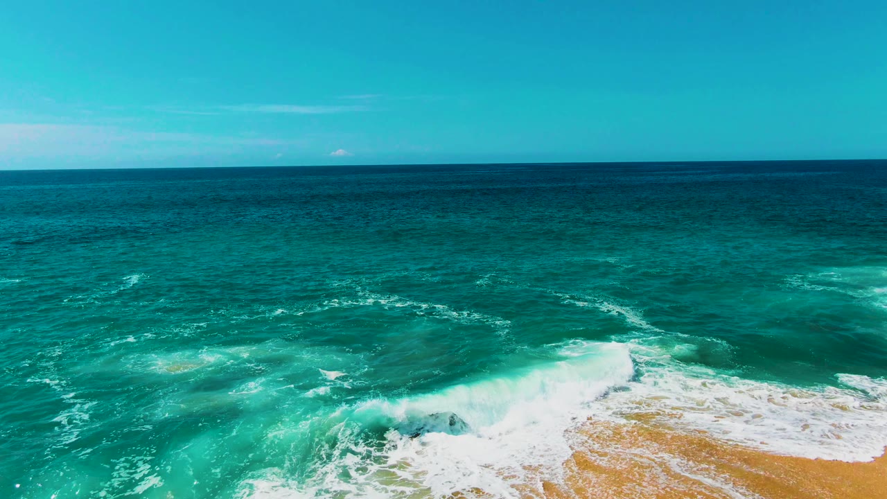 A second aerial moment at Punta Buzzards shows a broader band of white foam curling through turquoise water, with sand visible at the bottom right and no visible people.