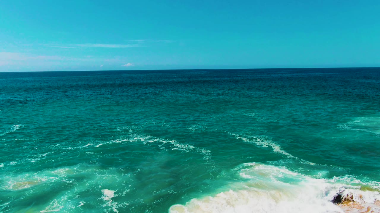 At Punta Buzzards, the shoreline shifts mostly out of view as turquoise water and white foam dominate the frame, with a small dark rock or reef patch near the lower right and no visible people.