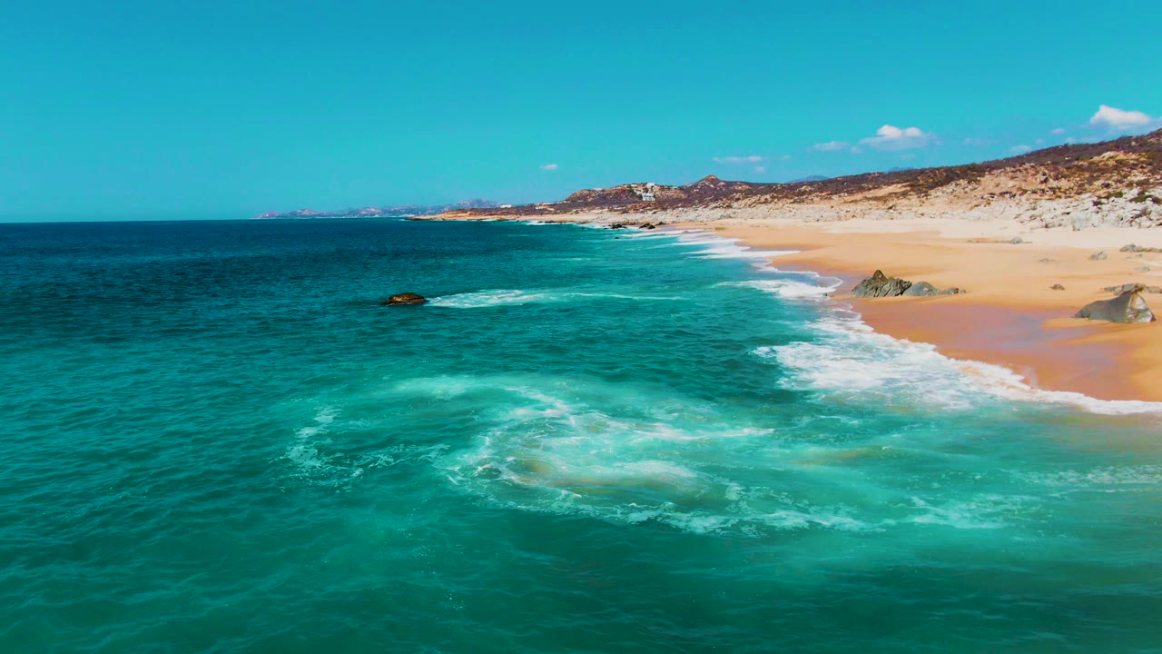 Aerial frame at Punta Buzzards shows a pale foam patch spreading in turquoise water, with the sandy beach and low scrub-covered hills beyond it, and no visible people.