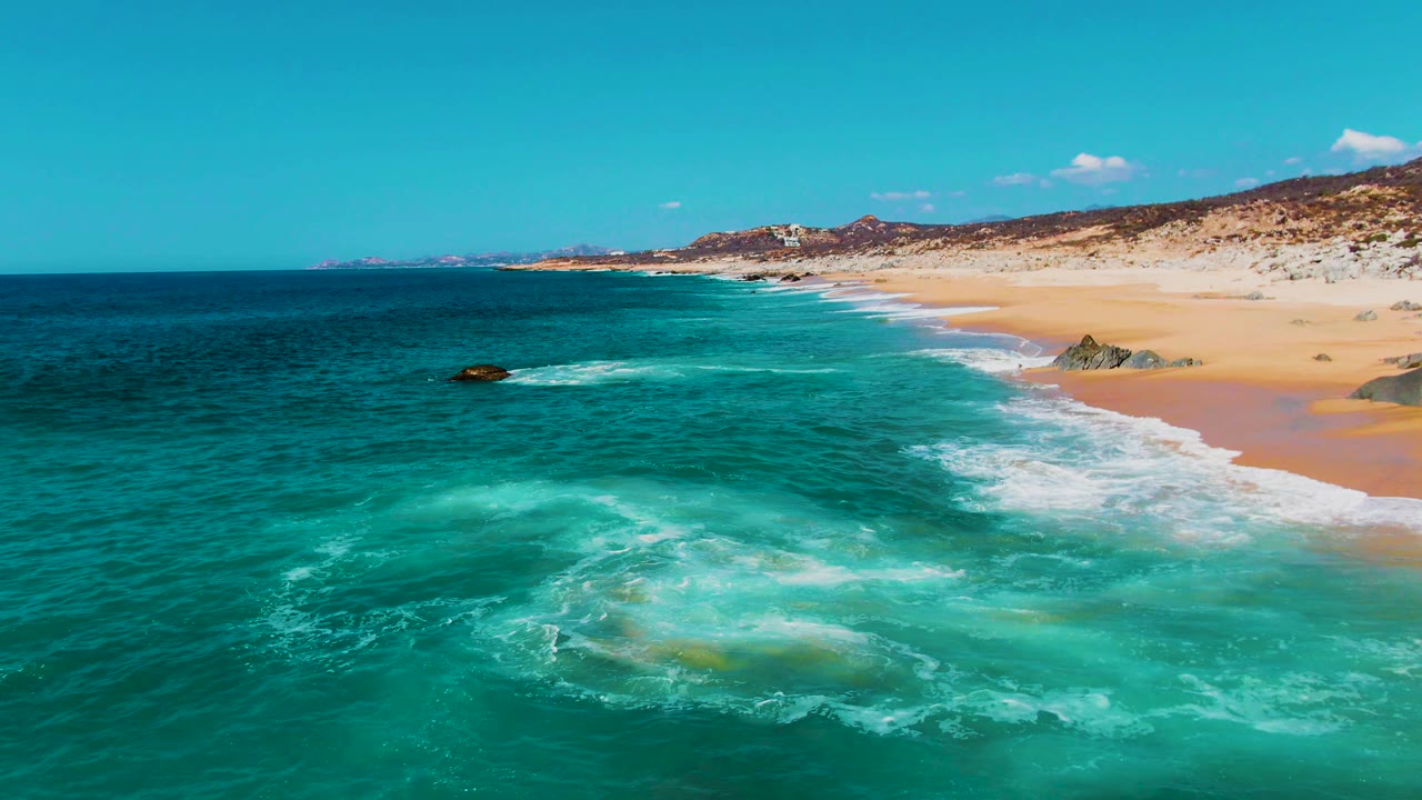 Punta Buzzards coastline appears from above with deeper blue water offshore, turquoise water near the breaking surf, tan sand, shoreline rocks, and no visible people.