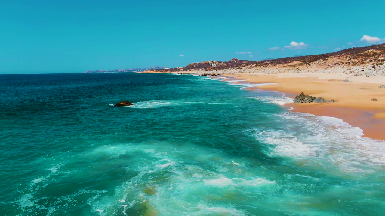 Aerial view over Punta Buzzards with white surf tracing the sandy beach, darker open water to the left, rocks near the shoreline, distant land on the horizon, and no visible people.