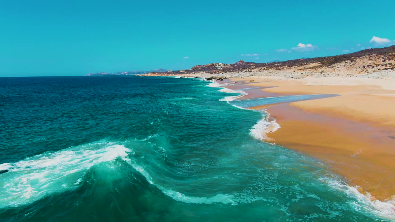 Turquoise surf breaking beside a wide sandy beach under a clear sky.
