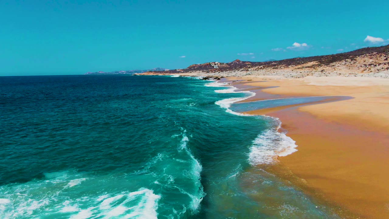 Aerial coastal frame from Buzzards Beach showing a foamy wave line meeting broad sand, deep blue water offshore, rocky terrain beyond the beach, and no visible people.