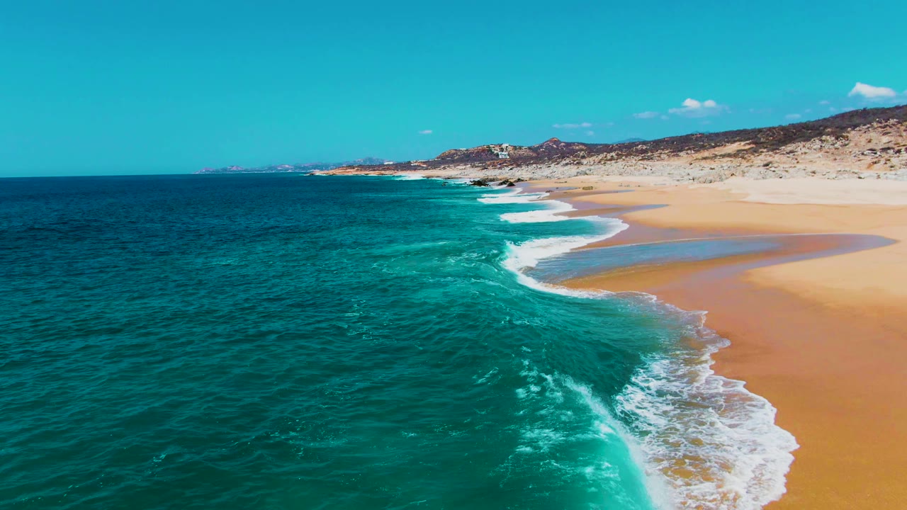 Drone-height view of Buzzards Beach in Baja California Sur with darker ocean water in the foreground, a bright surf break near the sand, rocky hills behind, and no visible people.