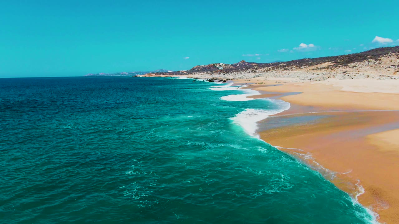 Aerial view along Buzzards Beach with a long curve of white surf, teal water filling the left foreground, pale sand to the right, rocky background hills, and no visible people.