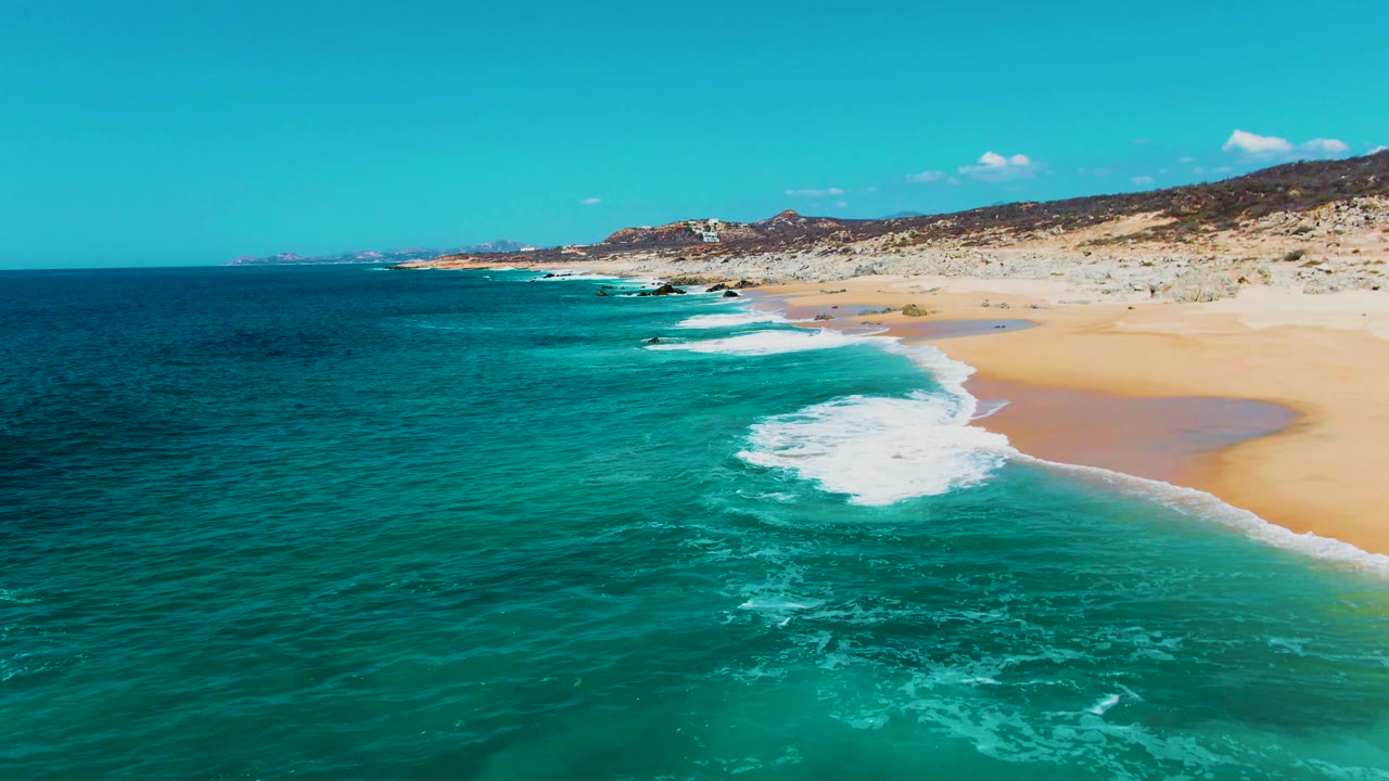 Aerial frame at Buzzards Beach with a larger patch of white foam spreading over turquoise water near the sand, rocky beach terrain beyond, and no visible people.