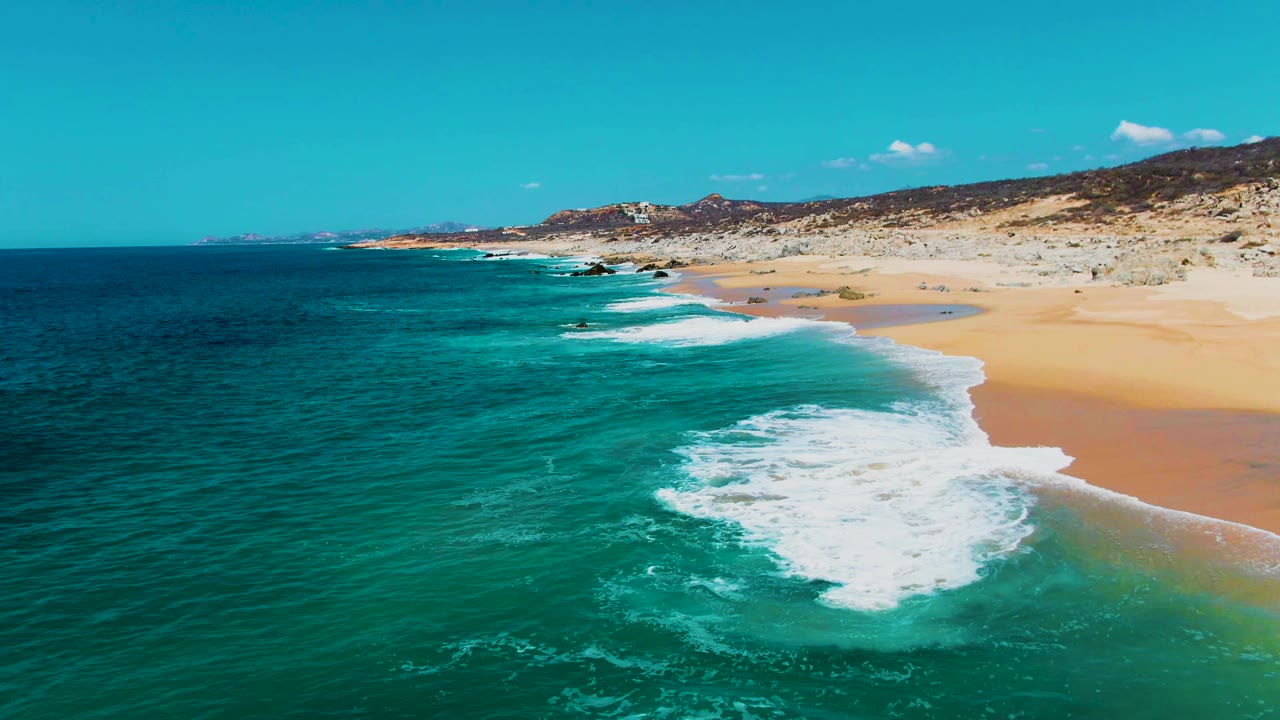 Aerial view of a breaking wave curling in turquoise water along Buzzards Beach, with a wide sand band to the right and no visible people.