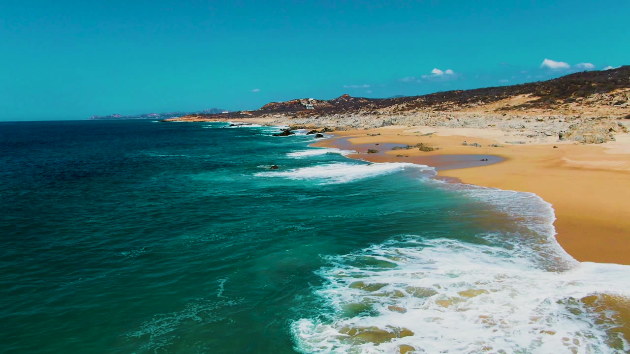 Aerial coastline view at Buzzards Beach showing surf lines washing onto sand, darker ocean water to the left, scattered rocks on the shore, and no visible people.