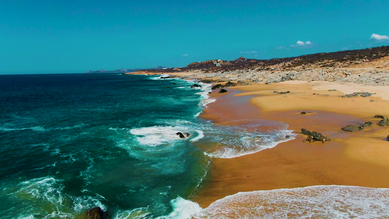 Aerial view of turquoise surf washing onto a sandy beach with dark rocks.