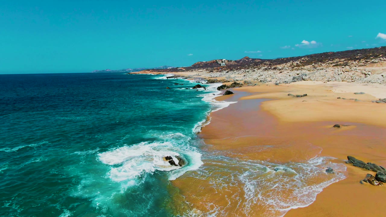 Aerial frame from Punta Buzzards, Baja California Sur, with a foamy wave wrapping around a dark rock, wet sand exposed on the beach, and no visible people.
