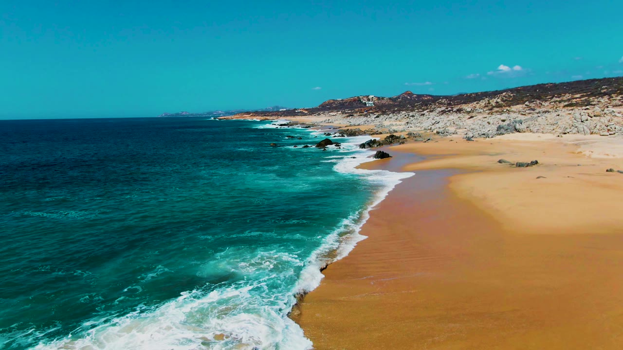 Aerial frame from Punta Buzzards, Baja California Sur, looking along a diagonal shoreline where turquoise water meets golden sand beneath rocky coastal terrain, with no visible people.