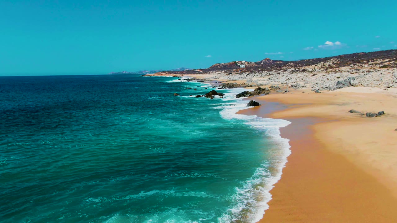 Aerial frame from Punta Buzzards, Baja California Sur, showing a broader stretch of blue ocean on the left, waves tracing the sandy beach, rocky outcrops, and no visible people.