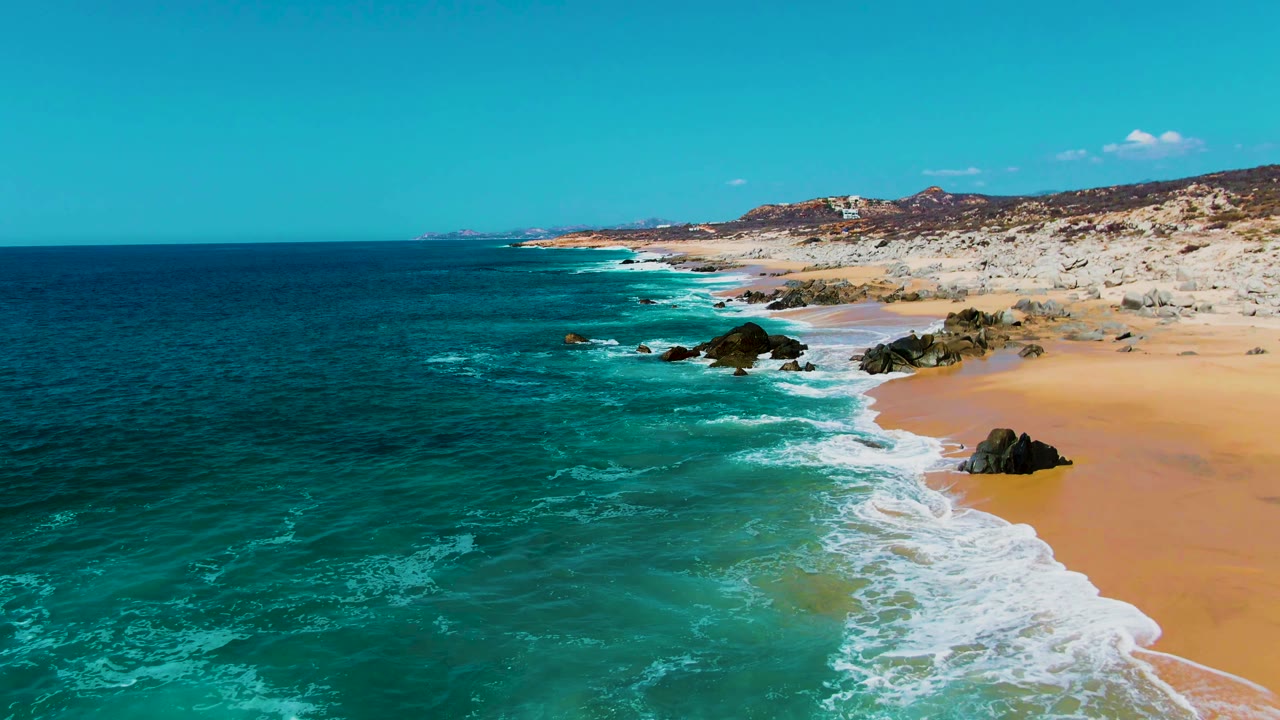 Aerial frame from Punta Buzzards, Baja California Sur showing turquoise ocean on the left, white surf crossing the sand, dark rocks near shore, pale dunes in back, and no visible people.
