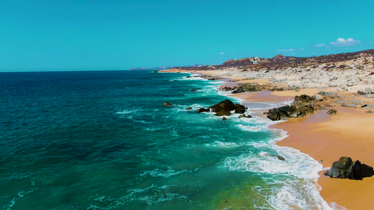Drone view of waves breaking around shoreline rocks on a sandy beach.
