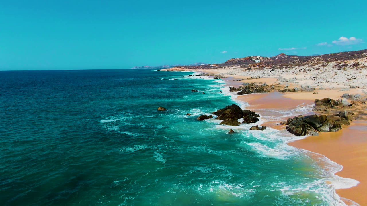 Aerial frame from Punta Buzzards, Baja California Sur looking along the coast as deeper blue water fills the left side, white surf curls toward rocks and sand on the right, with no visible people.
