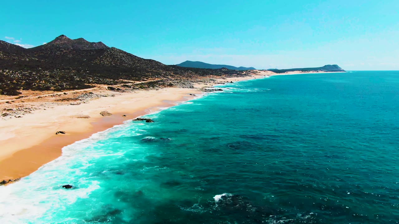 Aerial view of turquoise waves beside a sandy beach and dark mountains.