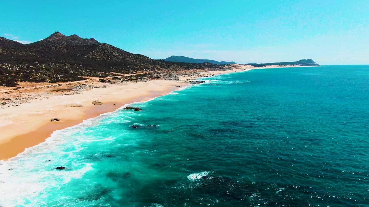 Aerial view at Punta Buzzards with white wave bands reaching the sandy shoreline, small dark rocks in the water, mountains on the left, and no visible people.