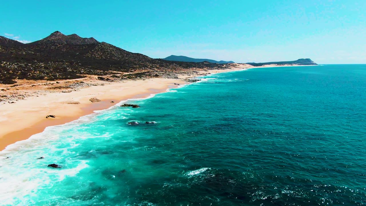 Punta Buzzards aerial frame with more open ocean visible on the right, turquoise shallows near the beach, a long sandy curve, mountain ridges inland, and no visible people.