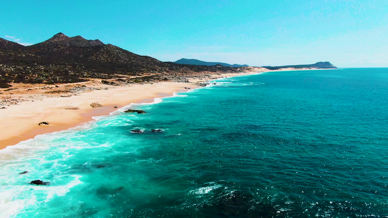 Mid-clip aerial view over Buzzards Beach with foamy surf tracing the sand, green-blue water in the foreground, scattered rocks near shore, mountains inland, and no visible people.