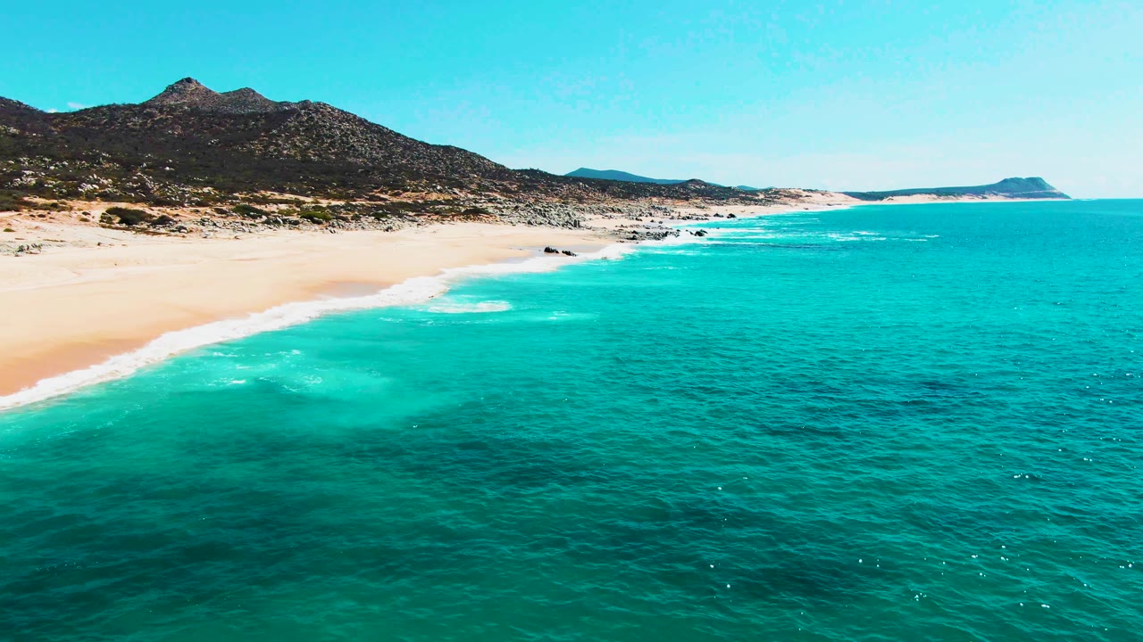 Aerial view of turquoise ocean beside sandy beach and dark mountains.