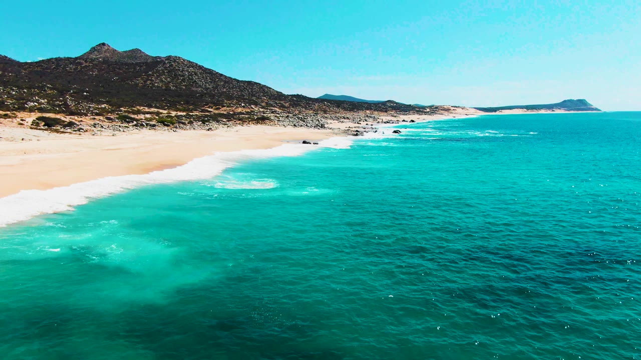 Aerial frame at Buzzards Beach showing a larger patch of turquoise water, white foam curling toward the sand, rocky coast in the distance, dark mountains, and no visible people.