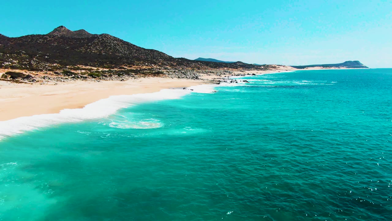 Aerial view at Punta Buzzards with the surf band stretched along the sand, clear blue-green water filling most of the frame, mountains on the left horizon, and no visible people.