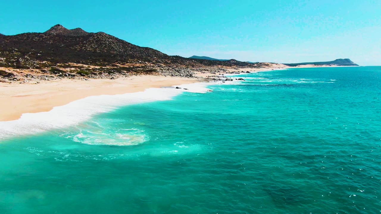 At Buzzards Beach, a bright foam patch spreads across shallow turquoise water near pale sand, with dark hills rising behind the coastline and no visible people.