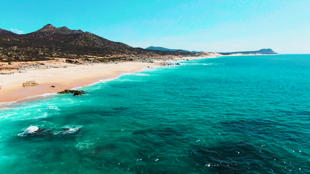 Opening frame of Punta Buzzards in Baja California Sur, with turquoise water in the foreground, pale beach and dark rocks along the shore, mountains behind, and no visible people.