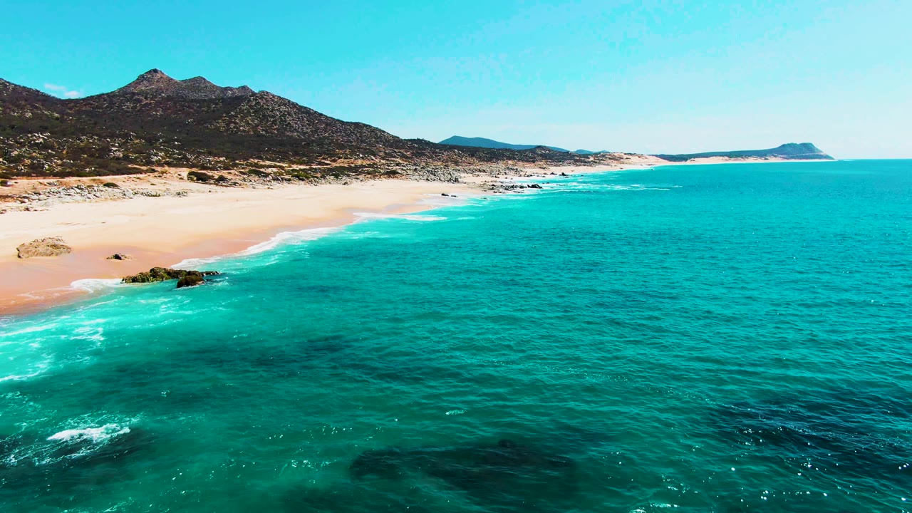 At one second, the Punta Buzzards shoreline shows gentle white surf tracking along the sand, dark reef patches under the water, mountains inland, and no visible people.