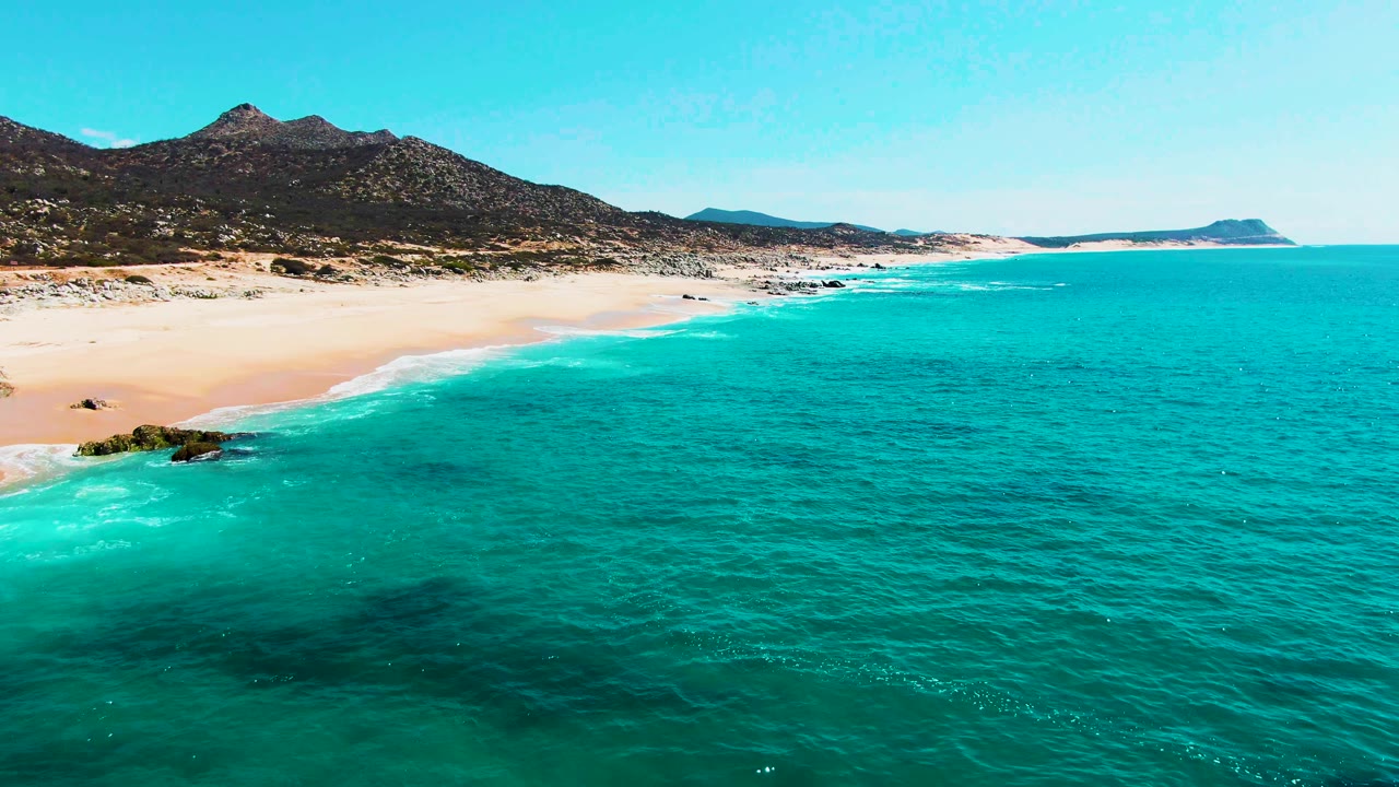 At two seconds, clear blue-green water fills more of the right side of the frame while the Punta Buzzards beach and rocky shoreline run toward distant mountains, with no visible people.