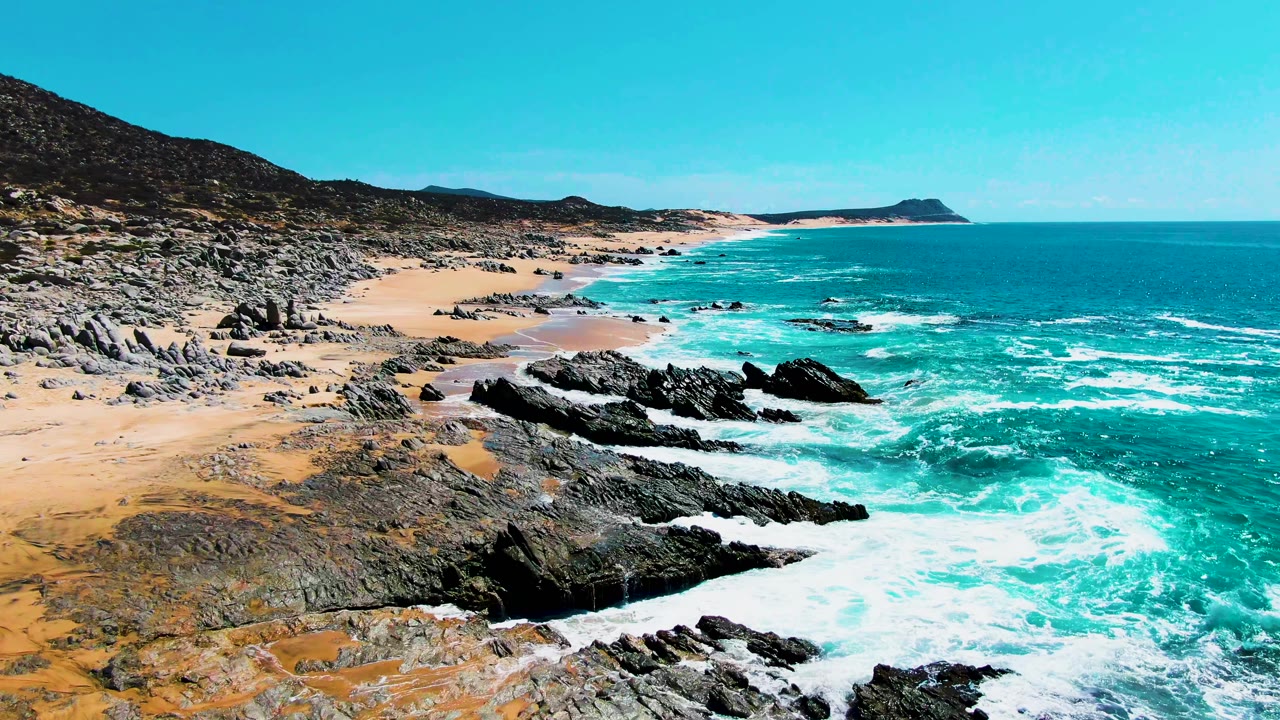 Aerial view of turquoise waves breaking around dark rocks on a sandy beach.