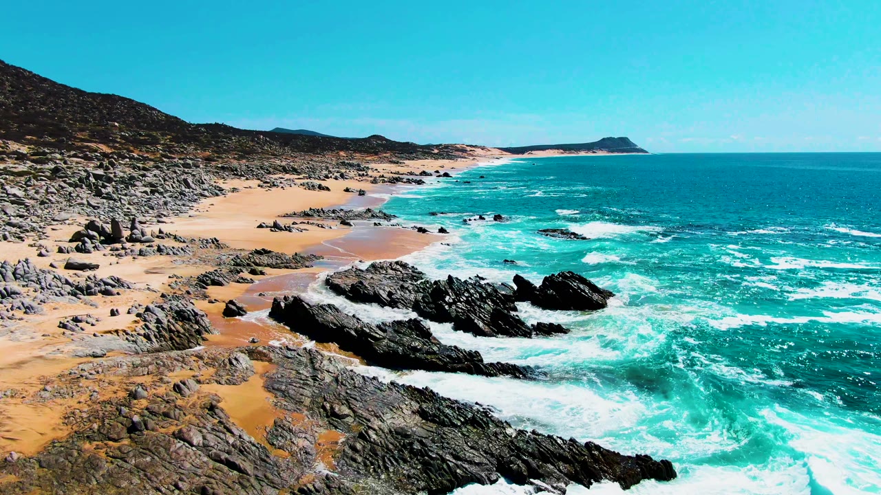 Aerial view at Punta Buzzards with surf wrapping around a central black rock formation, sandy coves to the left, open ocean to the right, and no visible people.
