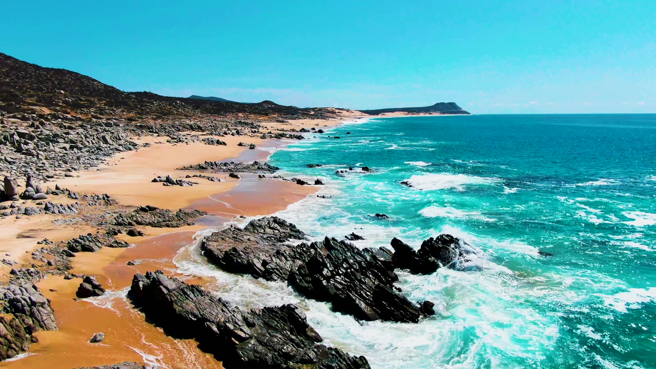 Aerial frame over Punta Buzzards where white water breaks hard against jagged foreground rocks, with empty sand, turquoise ocean, and no visible people.