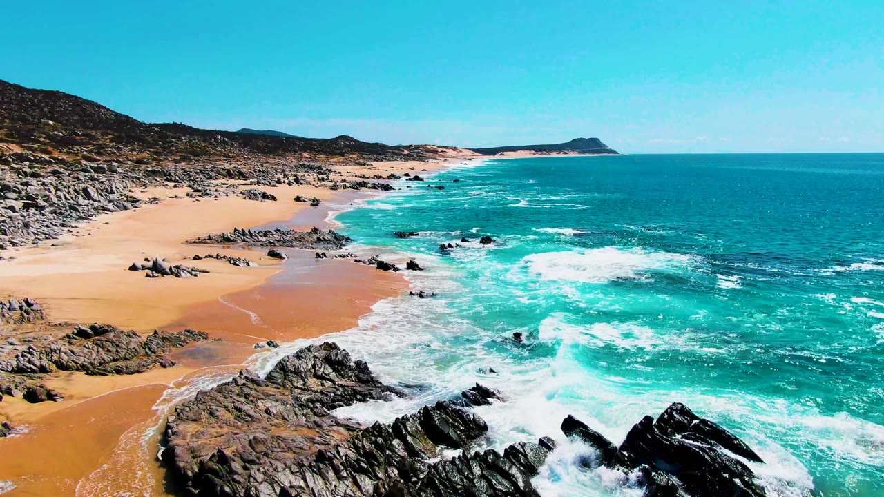 Wide aerial frame at Punta Buzzards showing a longer stretch of sandy shoreline, scattered rocks, white surf bands, blue water, distant headland, and no visible people.