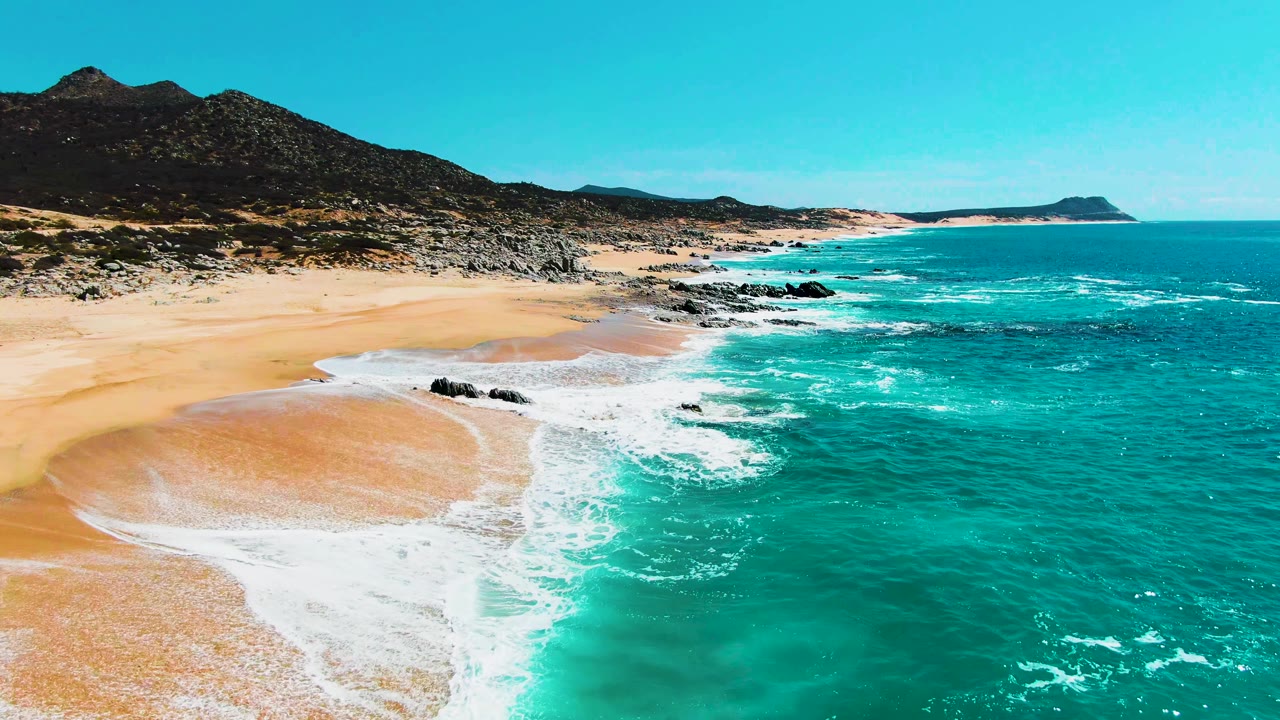 Aerial view of turquoise surf on a sandy beach with rocks and mountains.