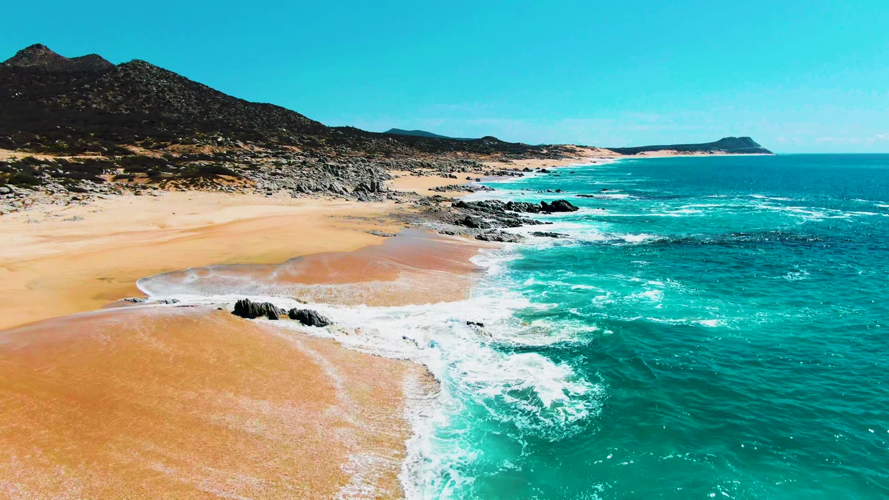 Aerial frame at Punta Buzzards, Baja California Sur, with white foam tracing the edge of wet sand, dark rock outcrops in the surf, mountains inland, and no visible people.